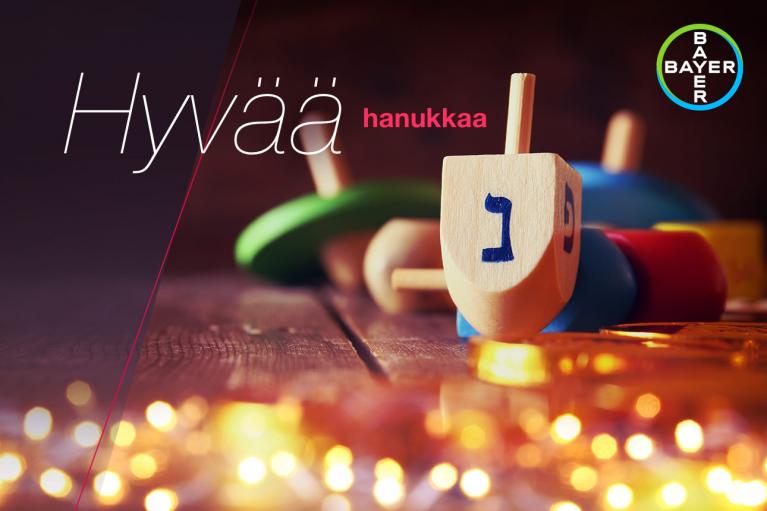 Wooden dreidel on a table with glowing lights, accompanied by the text "Hyvää hanukkaa" and a circular logo in the top right corner.