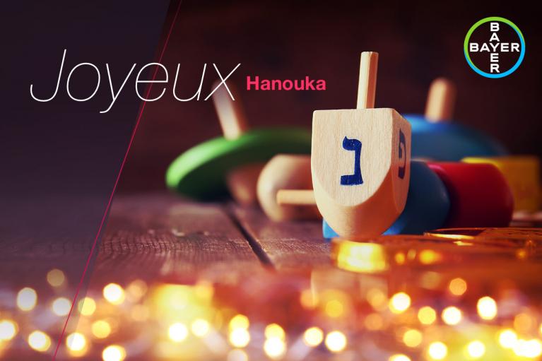 Wooden dreidel on a table with blurred lights in the foreground. The text reads "Joyeux Hanouka" and features a Bayer logo in the corner.