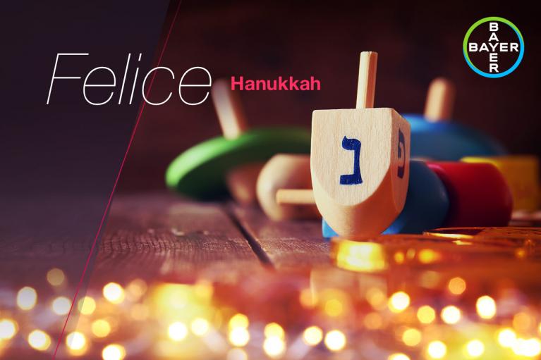 Wooden dreidels and gold coins on a table with a "Felice Hanukkah" greeting and Bayer logo in the corner.