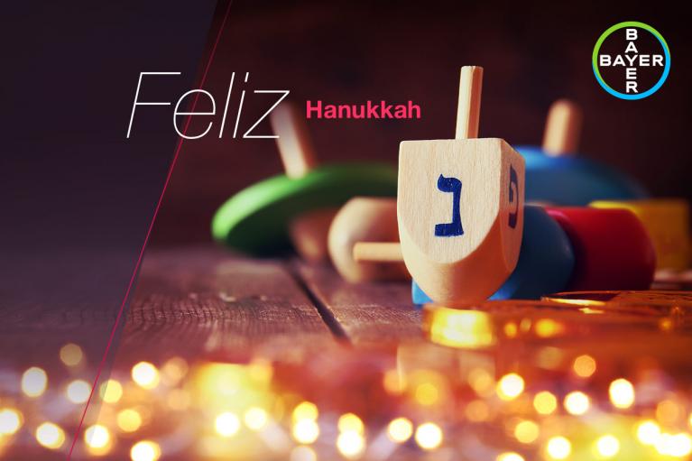 Colorful dreidels and coins on a wooden surface with the text "Feliz Hanukkah" and a Bayer logo in the corner.