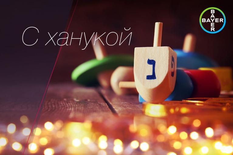 Wooden dreidel with Hebrew letter, surrounded by chocolate coins and colorful lights, featuring the Bayer logo, with Russian text wishing a happy Hanukkah.