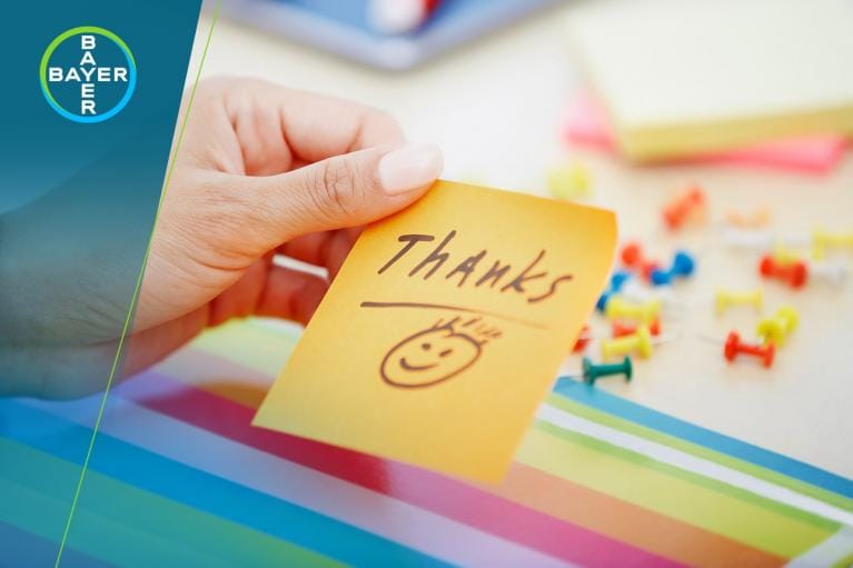 A hand holds a yellow sticky note with "Thanks" and a smiley face. Multicolored push pins and papers are visible in the background.