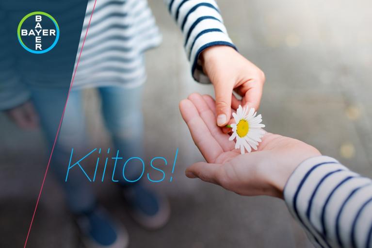 A person hands over two white daisies to another person. The word "Kiitos!" is written on the image. Bayer logo is visible in the corner.