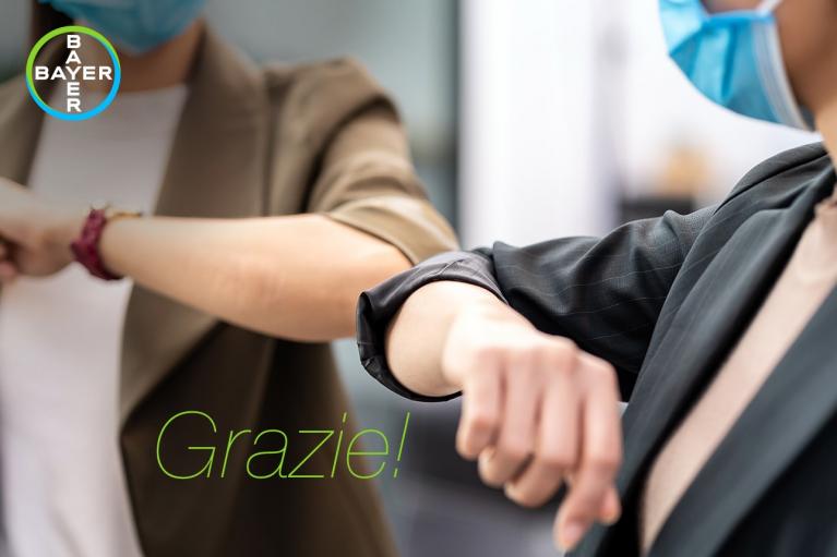 Two people wearing masks and blazers touch elbows. "Grazie!" is written below. The Bayer logo is in the top left corner.