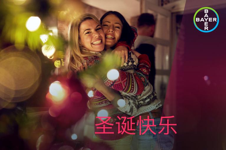 Two people in sweaters hug and smile near a decorated Christmas tree. Text says "圣诞快乐" with a Bayer logo in the corner.