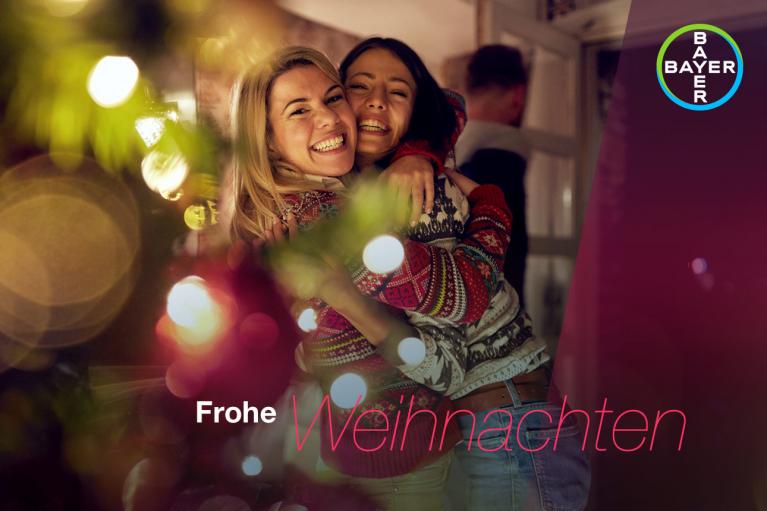 Two people hugging and smiling near a Christmas tree with the text "Frohe Weihnachten" and a Bayer logo in the corner.