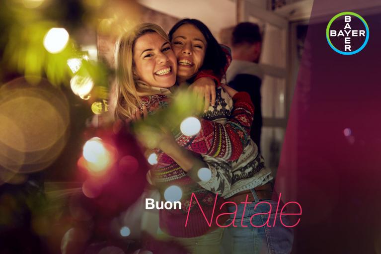 Two people smiling and embracing in front of a Christmas tree. The words "Buon Natale" and a Bayer logo are visible.