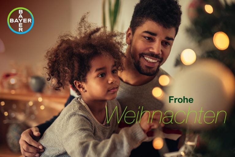 A man and child smile near a decorated Christmas tree. Text says "Frohe Weihnachten" and the Bayer logo is visible.