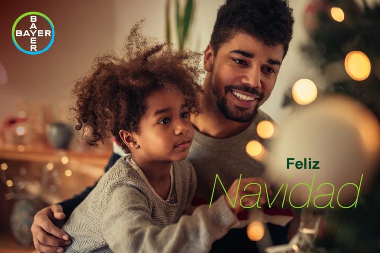 A man and child decorate a Christmas tree. Text reads "Feliz Navidad" with a Bayer logo in the corner.