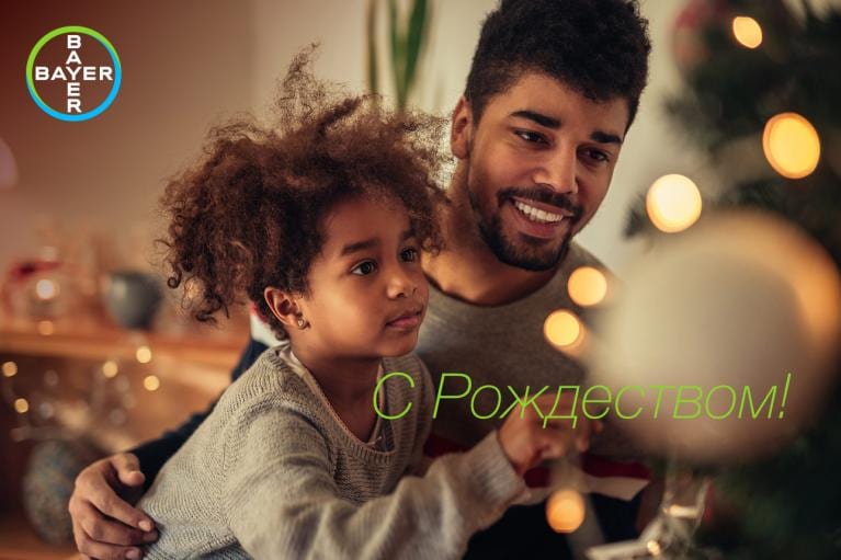 A man and a child decorate a Christmas tree with lights and ornaments. The phrase "С Рождеством!" is displayed, and the Bayer logo is present in the corner.