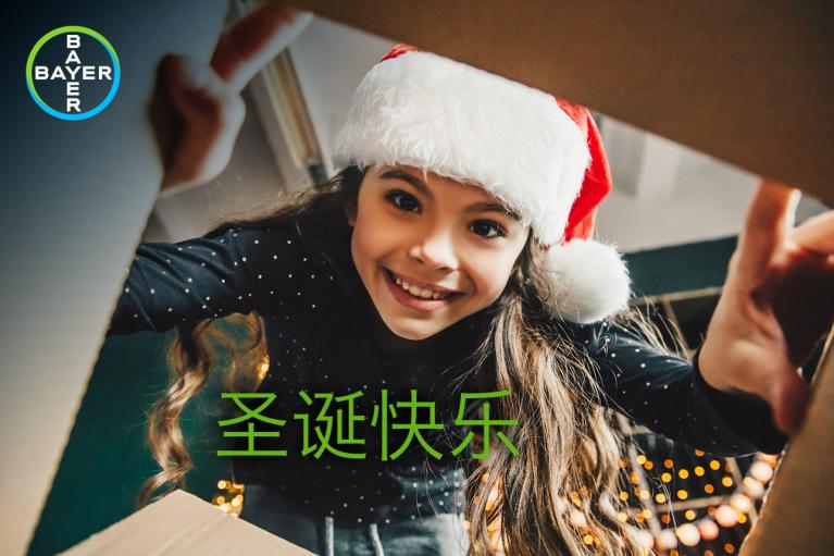 Girl wearing a Santa hat smiles through a cardboard box with Chinese text for "Merry Christmas." Bayer logo in the upper left corner.