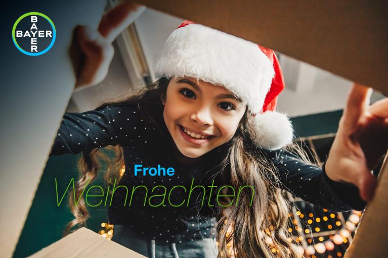 Girl wearing a Santa hat smiles inside a cardboard box with the text "Frohe Weihnachten." Bayer logo in the top left corner. Holiday lights in the background.