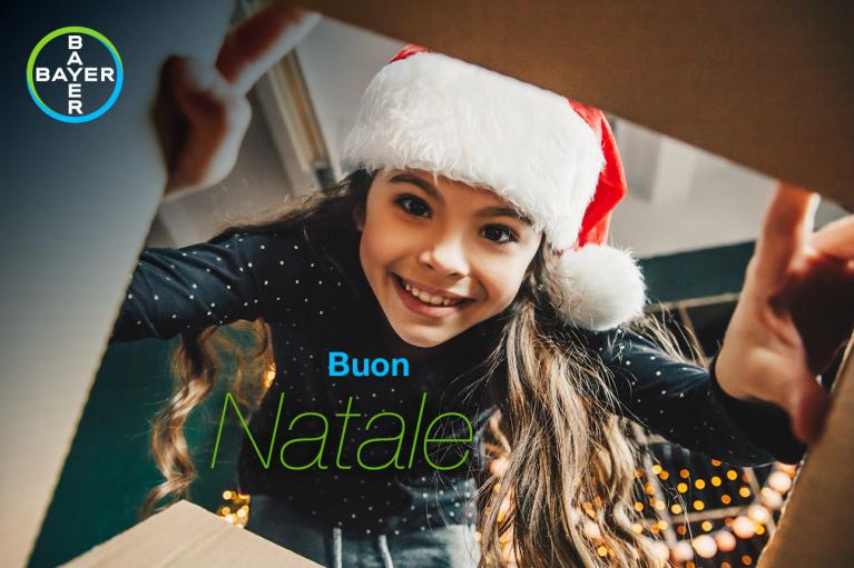 A young girl in a Santa hat looks into a cardboard box with "Buon Natale" text and a Bayer logo in the corner.
