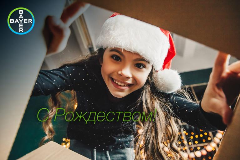 A girl wearing a Santa hat looks into a box, with "С Рождеством" written in green letters. The Bayer logo is in the top left corner.