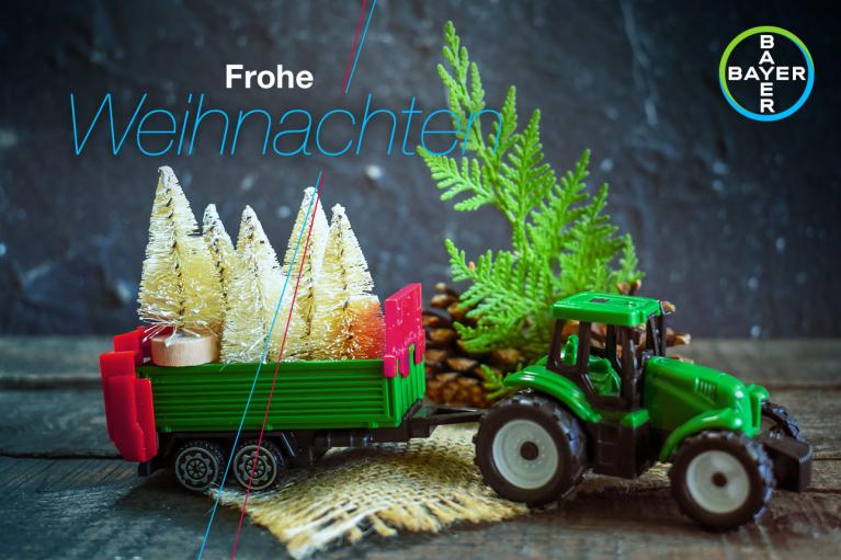 Toy tractor hauling miniature Christmas trees with the text "Frohe Weihnachten" and the Bayer logo.