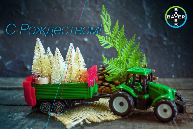 A small green toy tractor pulls a trailer with miniature Christmas trees. A pine cone and evergreen sprig are in the background. Text in Russian reads, "С Рождеством!" and a circular logo is in the corner.