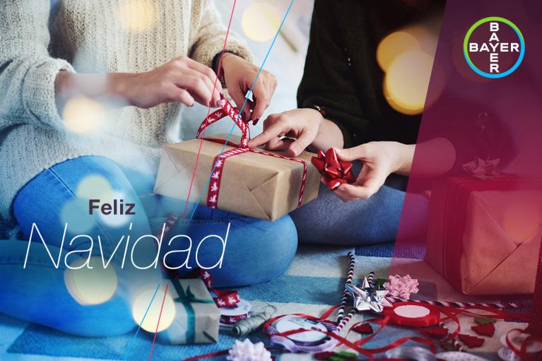 Two people wrapping a gift with red ribbon, surrounded by festive wrapping materials. Text reads "Feliz Navidad" with a Bayer logo in the corner.