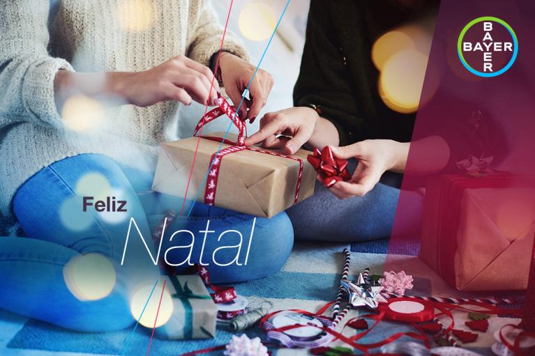 Two people wrapping a gift with red ribbon on a blanket. The words "Feliz Natal" are overlaid, alongside the Bayer logo.