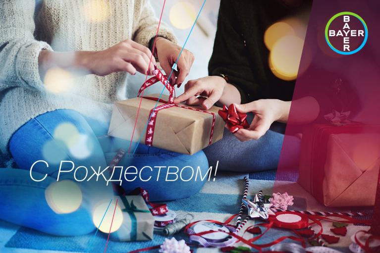 Two people wrapping a gift with red ribbon, surrounded by wrapping supplies. Text in Cyrillic reads "С Рождеством!" and a Bayer logo is visible in the corner.