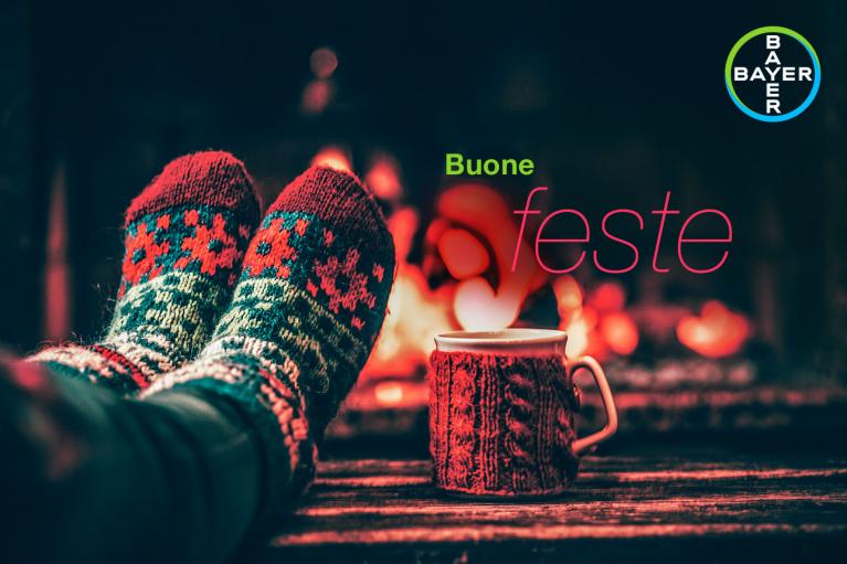 Cozy scene with feet in knitted socks near a warm fire and a patterned mug, featuring the text "Buone feste" and Bayer logo.