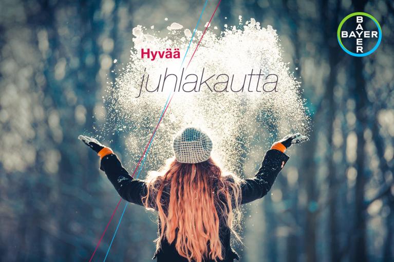 Person in winter attire stands with outstretched arms as snow is tossed in the air; text reads "Hyvää juhlakautta" with the Bayer logo in the corner.