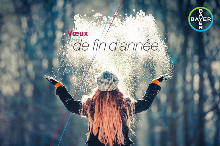 Person in winter clothing joyfully throws snow in the air in a forest. Text reads, "Vœux de fin d'année," with a Bayer logo in the corner.