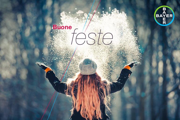 Person in winter clothes with arms raised, facing falling snow, with the text "Buone feste" and Bayer logo in the corner.