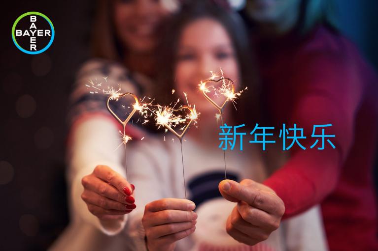 Three people holding sparklers shaped like hearts with the text "新年快乐" and a Bayer logo in the corner.
