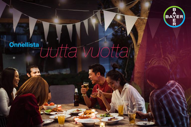 Group of people laughing and enjoying a meal at a table outdoors with festive string lights and a "Happy New Year" message in Finnish.