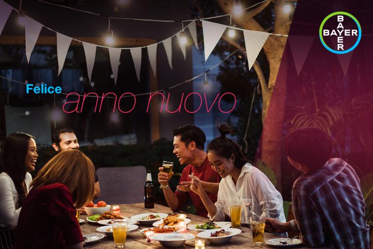 A group of people enjoying a meal outdoors under string lights with the words "Felice anno nuovo" in the background.
