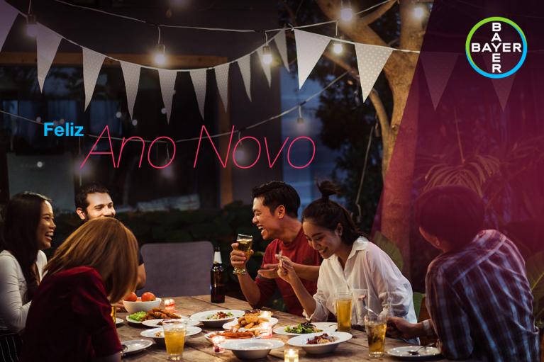 A group of people sits around an outdoor table with food and drinks, celebrating under string lights and banner flags. "Feliz Ano Novo" is in the background with a Bayer logo.