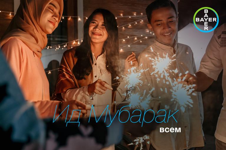 Three people smiling and holding sparklers while celebrating. Strings of lights are in the background. Text on the image reads, “Eid Mubarak всем.”.