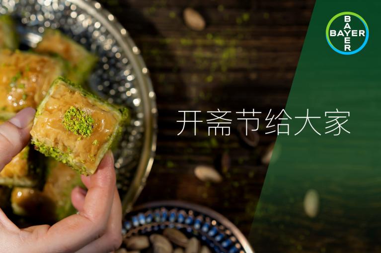 Close-up of a person holding pistachio baklava. A decorative plate is visible, and the Bayer logo appears in the corner with Chinese text overlay on the image.