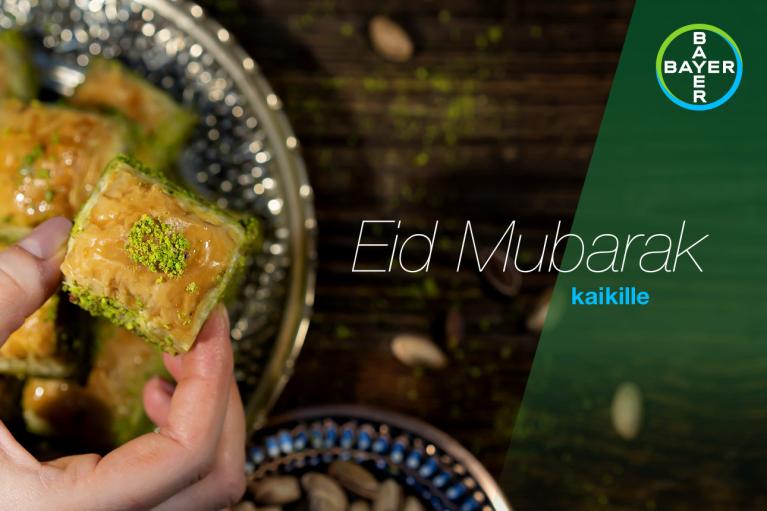 A hand holding a piece of baklava with a "Eid Mubarak kaikille" message and a Bayer logo in the background.
