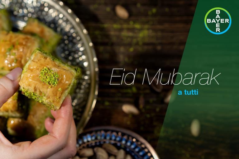 A hand holds a piece of pistachio baklava over a plate. Text reads "Eid Mubarak a tutti" with a Bayer logo in the corner.