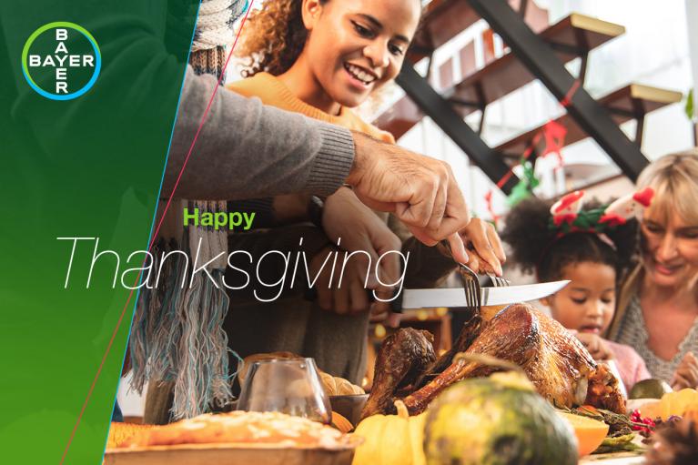 Family gathers around dining table with Thanksgiving meal, including a turkey, as a person carves. "Happy Thanksgiving" text and Bayer logo are visible.