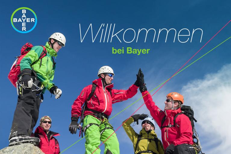 Group of climbers in colorful gear high-fiving on a mountain with a clear blue sky. Text reads "Willkommen bei Bayer." Bayer logo is visible.