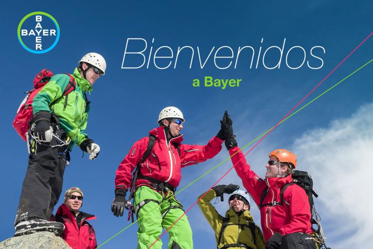 Group of climbers in colorful gear celebrating atop a peak, beneath a "Bienvenidos a Bayer" sign.