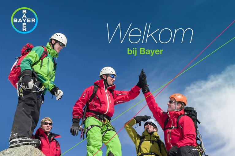 Four climbers wearing helmets and colorful gear celebrate on a mountain peak under a clear blue sky with a "Welkom bij Bayer" message.