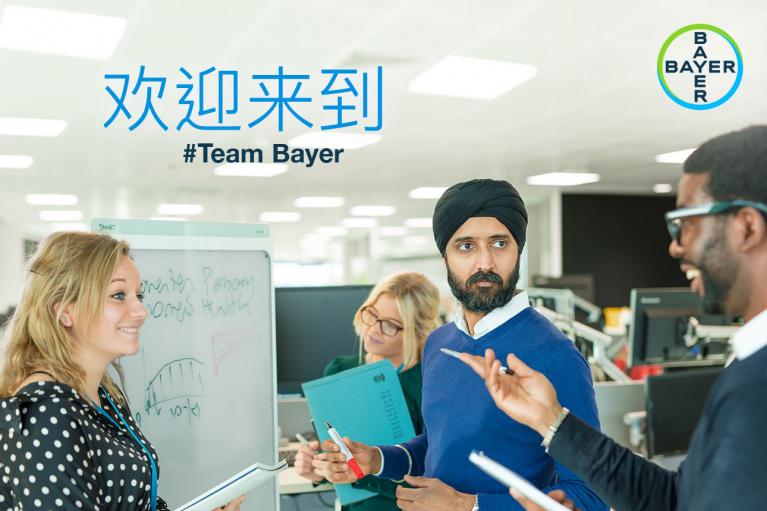 A diverse group of four colleagues engaged in discussion around a whiteboard in an office, with a Bayer logo and Chinese text reading "#Team Bayer" above them.