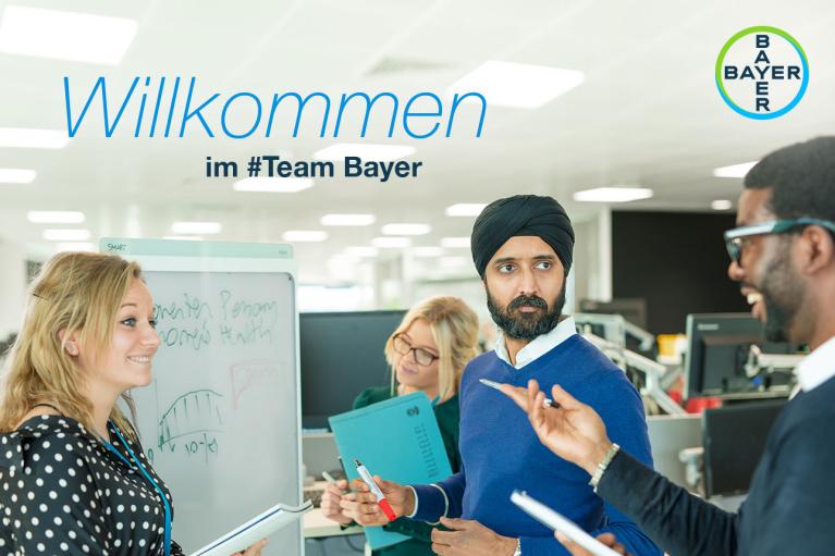 A diverse group of four people collaborating around a whiteboard in an office. Text reads "Willkommen im #Team Bayer." Bayer logo is visible.