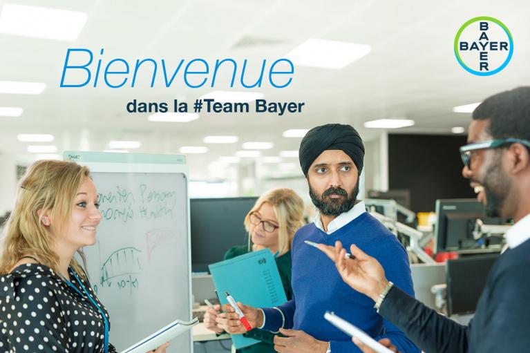 Four professionals in a discussion at an office, with a whiteboard in the background and the text "Bienvenue dans la #Team Bayer" above.