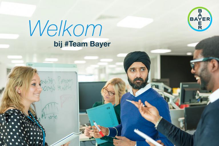 Four colleagues engage in a discussion at an office. A whiteboard with a graph is in view. The text reads "Welkom bij #Team Bayer," accompanied by the Bayer logo.