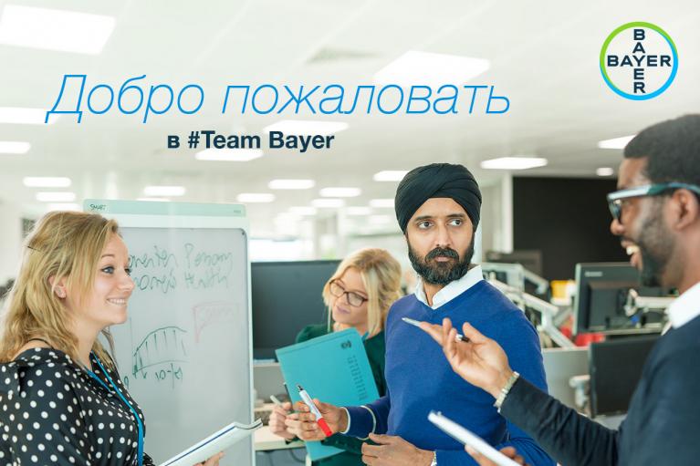 Group of four colleagues discussing around a whiteboard in an office. The text says "Добро пожаловать в #Team Bayer" with Bayer logos in the image.