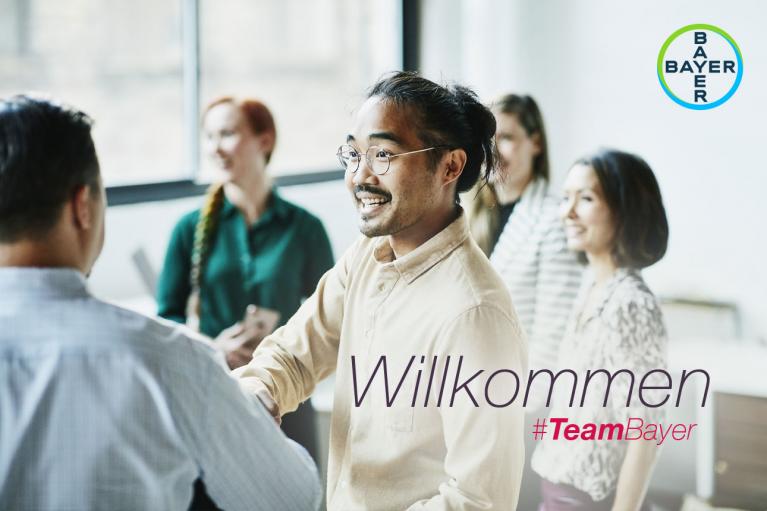A group of people in an office setting, one man shaking hands. Text reads "Willkommen #TeamBayer." Bayer logo is visible.