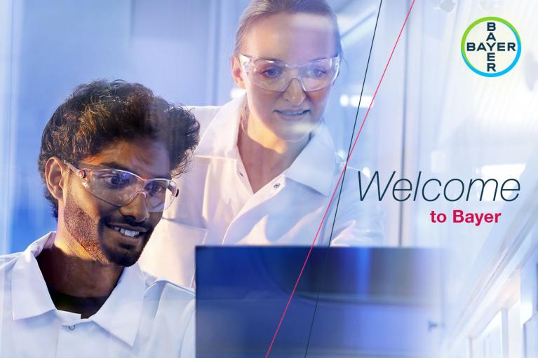 Two scientists in lab coats and safety goggles work together in a lab. A Bayer logo and the text "Welcome to Bayer" are visible.