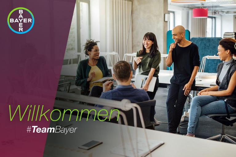 Group of five colleagues smiling and talking in a modern office setting with "Willkommen #TeamBayer" written on a banner.