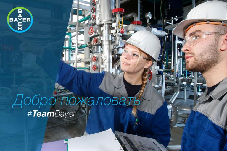 Two workers in safety gear discuss operations in an industrial facility. One points to equipment while holding a clipboard. Bayer logo and the phrase "Добро пожаловать #TeamBayer" are visible.