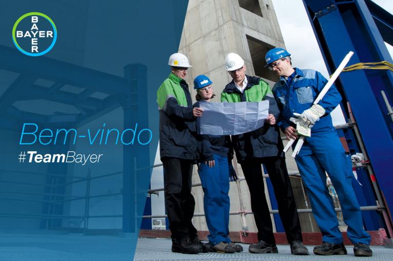 Four construction workers in safety gear examine a blueprint at a building site. Bayer logo and "Bem-vindo #TeamBayer" text are visible.