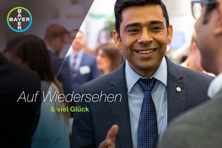 A person in a suit smiles during a business event with a Bayer logo and German text reading "Auf Wiedersehen & viel Glück" on the image.